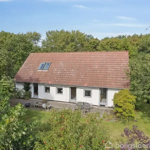 Hus med rødt tegltag står midt i frodig have med tætte træer. Terrasse med havemøbler foran huset. Klar himmel danner baggrund.