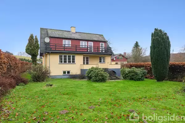 Et to-etagers hus med gul facade og røde detaljer står i en have med grøn plæne og buske. Træer og en hæk omgiver huset, der har en balkon på øverste etage.