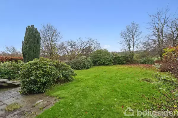 En velplejet græsplæne, omgivet af buske og træer, ligger i en have. En smal flisebelagt sti ses i venstre side. Himlen er klar og blå.