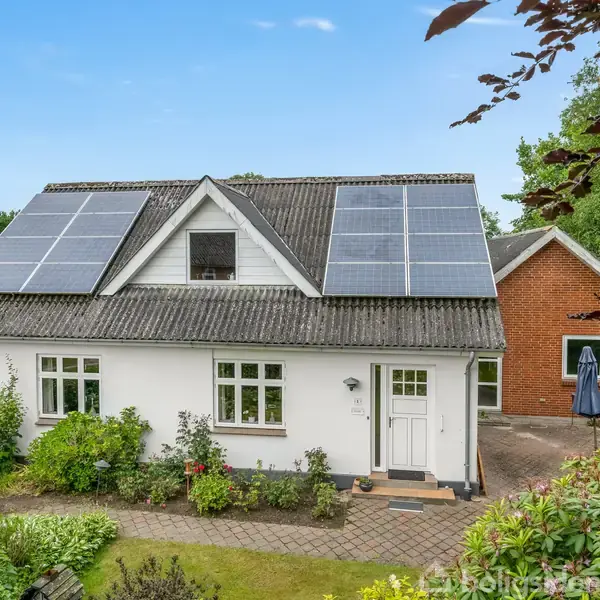 Et hvidt hus med solpaneler på taget står blandt frodige grønne buske og træer. Til venstre ses en velplejet have, og til højre en rød bygning.
