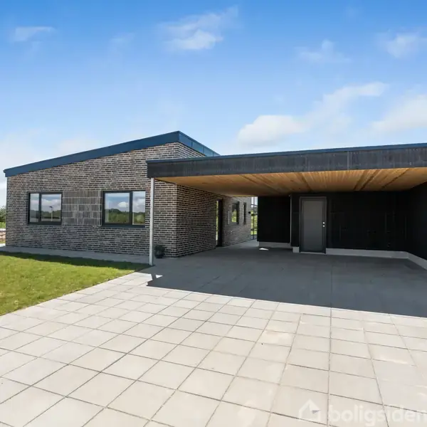 Et moderne hus i mursten står stille ved siden af en carport, omgivet af en flisebelagt indkørsel og en grøn græsplæne under en blå himmel.