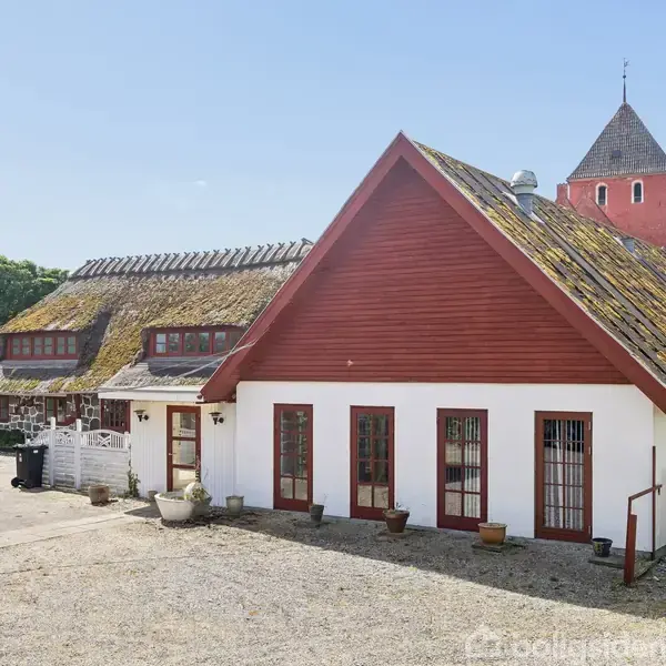 Et traditionelt bindingsværkshus med rødt tag og hvide vægge står på en grusvej. Omkring huset er der frodig vegetation og en kirketårn kan ses i baggrunden.