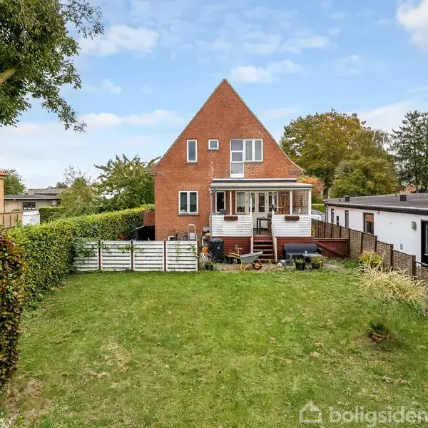 Rødt murstenshus med saddeltag står centralt. Foran huset er en grøn græsplæne omgivet af hække; en terrasse med trapper ses foran huset. Himmelen er blå med få skyer.