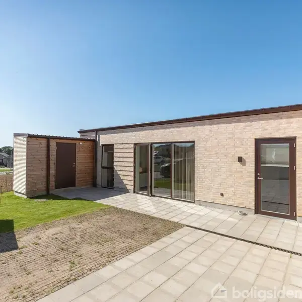 Et moderne hus med store glasdøre står stille i en solrig, flisebelagt terrasseomgivelser. Til venstre ses en lille græsplæne og en træbeklædt skur i et villakvarter.