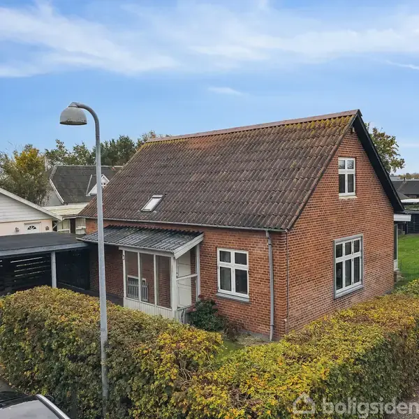 Rødt murstenshus med skråt tag og hvide vinduer står på en parcel med hæk omkring. Ved siden af huset er en carport. En gadelampe ses foran i billedet.