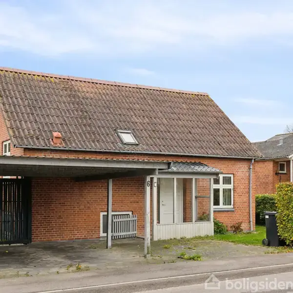 Et rødt murstenshus står langs en vej med tag af røde tegl og en carport til venstre. En bænk er placeret ved en hvid dør, omgivet af buske og en skraldespand.