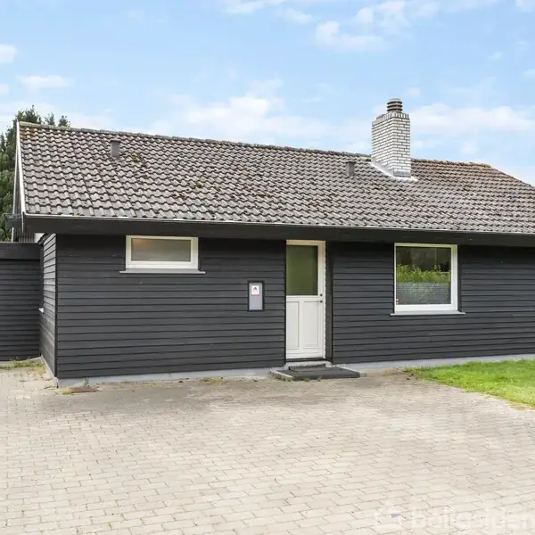 Sort hus med skråt tag og skorsten, står stationært. Huset er omgivet af en flisebelagt indkørsel og græsplæne med træer i baggrunden.