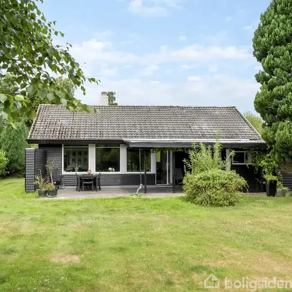 Et sort hus med skråt tag står stille i en grøn have, omgivet af store træer og buske på en overskyet dag.