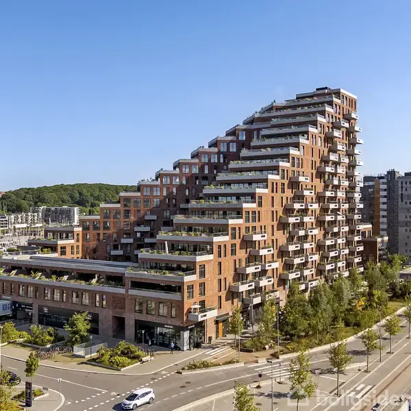 Et terrasseret boligtårn rejser sig trappevis med grønne altaner. Bygningen ligger langs en bred vej ved en kanal og er omgivet af træer og moderne arkitektur.