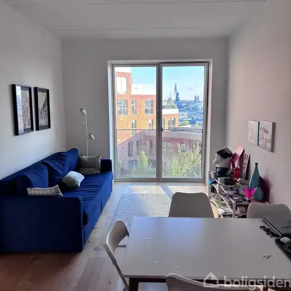 En blå sofa står i en stue mod en væg med billeder. Store glasdøre fører ud til en udsigt over bygningskomplekser. Et bord med stole er i forgrunden.