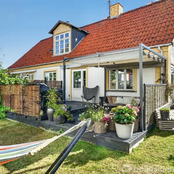 Et hus med rødt tegltag har en terrasse med blomsterkrukker og en sort stol. En farverig hængekøje hænger i haven, omgivet af grønne buske og græsplæne.