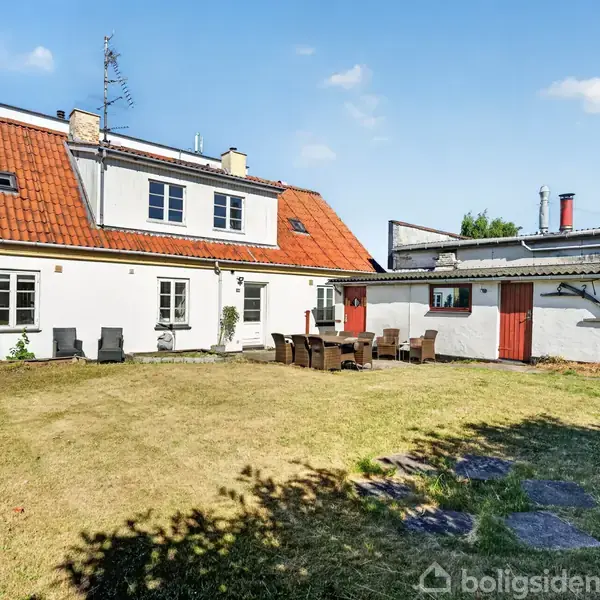 Hvidt hus med rødt tag står i en have. Foran huset er et bord og stole, omgivet af en græsplæne under en klar, blå himmel.