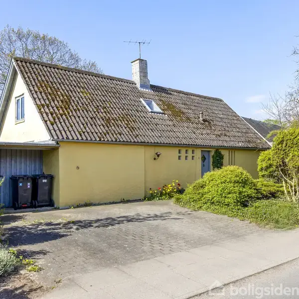 Gul hus med skråtag har to skraldespande ved siden af en carport. Omgivet af buske og træer på en stille vej under en blå himmel.