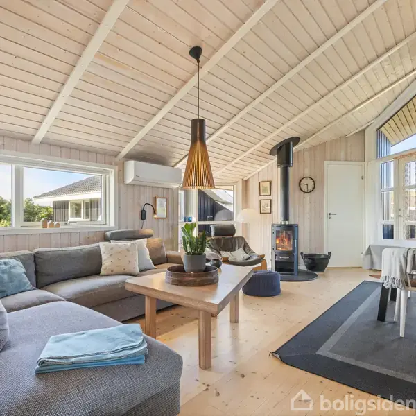 Stue med grå hjørnesofa, sofabord og pejseovn i hjørnet. Spisebord med stole ved gulvtæppe. Trævægge og loft, store vinduer giver lysindfald. Moderne, hyggelig atmosfære.