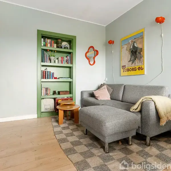 Grå sofa placerer sig i hjørne med tæppe og puder, omgivet af bøger i en grøn reol. Væggene har plakater, og et orange spejl hænger til pynt. Trægulve.