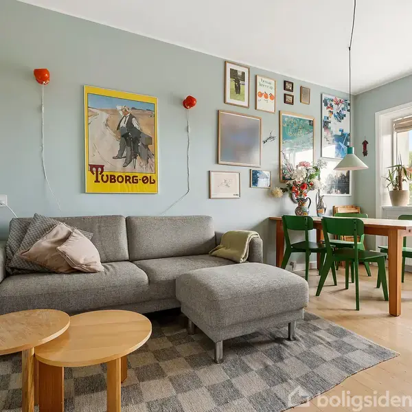 Grå sofa med puder, stående på tæppe, omgivet af kunst på væggen. To runde borde foran sofaen. Spisebord med grønne stole ved vindue. Tekst: "Tuborg-øl."