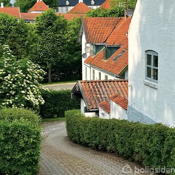 Et lyst køkken med hvide skabe og en træbordplade. Køkkenet har åbne hylder med service, en opvaskemaskine, og store vinduer, der giver udsigt til en terrasse med planter.