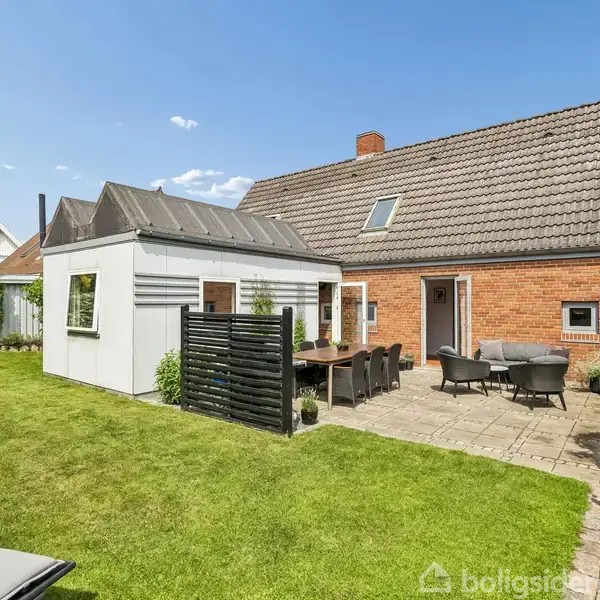 Et rødt murstenshus med et sort tegltag, står ved siden af en grøn have. Foran huset er der en terrasse med havemøbler, omkranset af hække og græs.