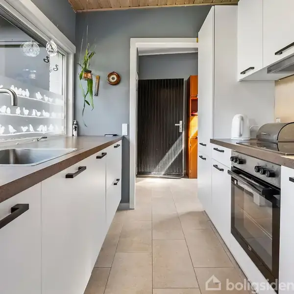 Et moderne køkken med hvide skabe og en brun bordplade. En håndvask findes ved vinduet, mens en ovn og kogeplade er til højre. Det er sollys udenfor.