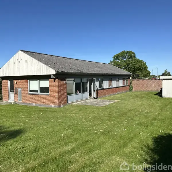En enkeltstående, muret etplanshus med røde mursten og tag i sort tegl står på en velholdt græsplæne i et grøn haveområde med en blå himmel som baggrund.
