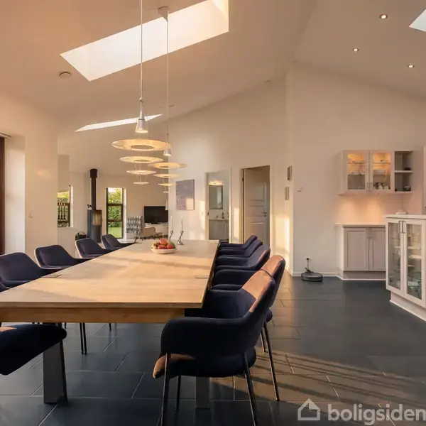 Stor spisebord omgivet af blå stole i en lys spisestue med ovenlys og glasdøre til haven. Moderne loftslamper hænger over bordet, og køkkenet er synligt for enden.