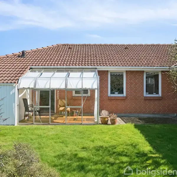 En rød murstensbungalow med glasoverdækket terrasse, beliggende i en grøn have med blå himmel i baggrunden. Terrassen har stole og bord, omgivet af træer og buske.