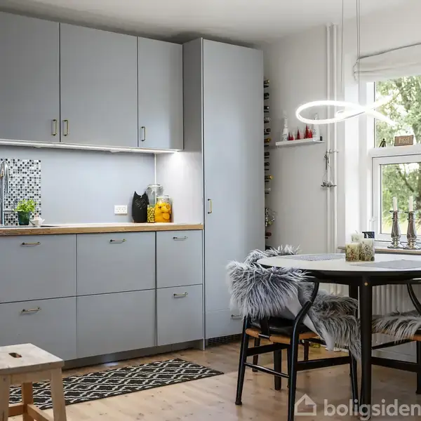 Et moderne køkken med lysegrå skabe står langs en væg. Et lille spisebord med stole beklædt med pels er under en cirkulær lampe. Store vinduer slipper naturligt lys ind.