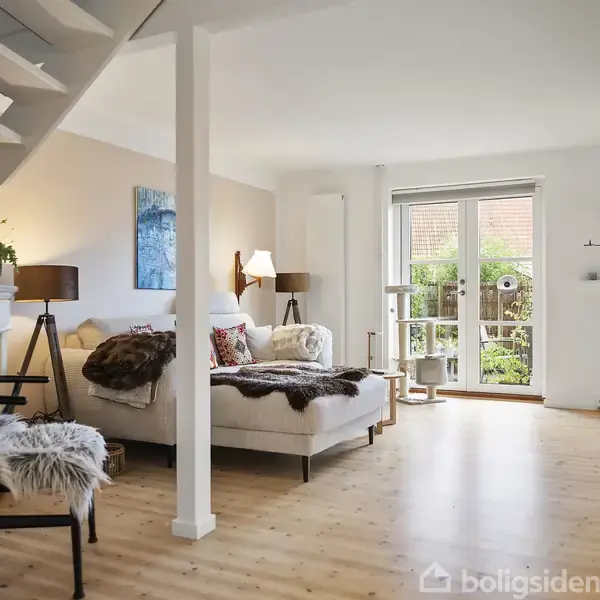 Stue med sofa, puder og tæpper står i hjørnet. Reol og lampe ved siden af; vinduer viser terrasse med planter. Trappe fører opad. Indretningen er lys og moderne.