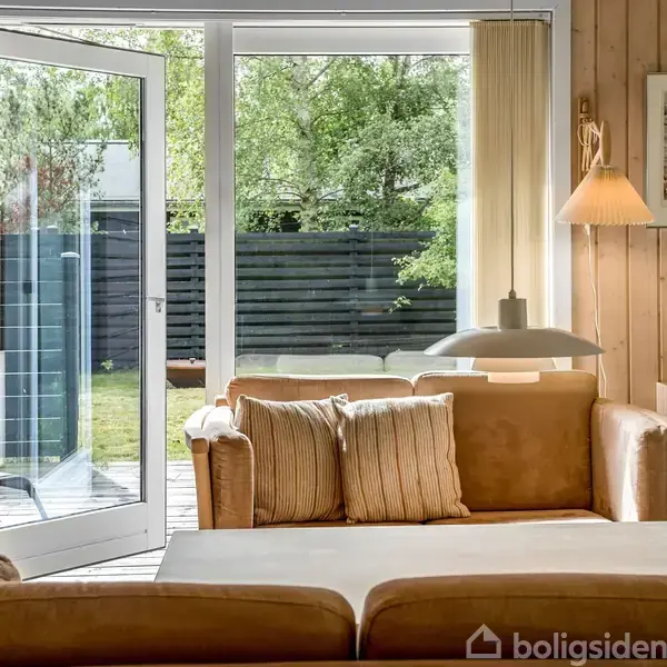 En lys stue med en lædersofa nær en åben havedør. Sollys strømmer ind, mens puder ligger på sofaen. I baggrunden er der trævægge, en lampe og et maleri.