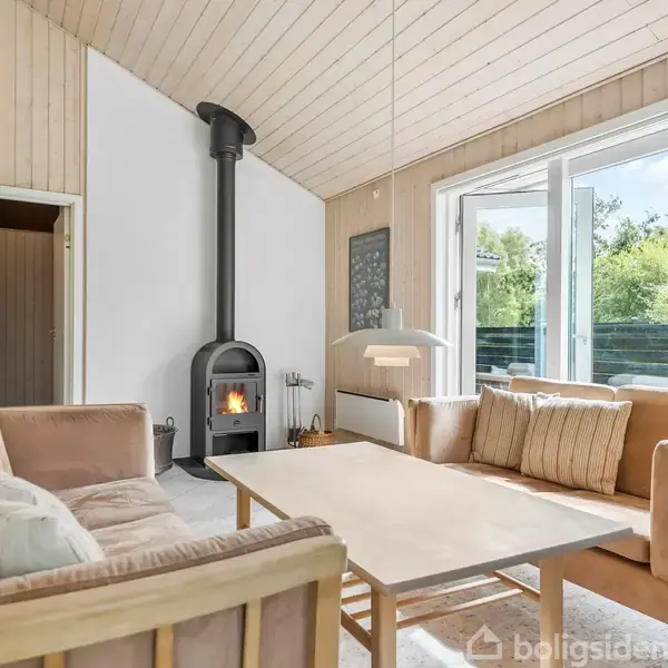 En hyggelig dagligstue med en brændeovn, der står ved væggen og varmer rummet. To sofaer omkranser et sofabord. Store vinduer slipper masser af lys ind.