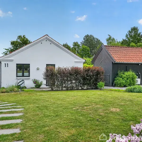 Et hvidt hus med nummer 11 står stille i en frodig have med grøn plæne og buske, omgivet af farverige blomster og et mindre anneks.
