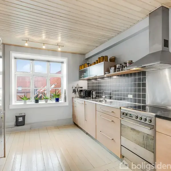 Et moderne køkken med rustfrit stål-apparater og lyse træskabe. Der er en stor vindue med udsigt, planter og diverse køkkenredskaber på hylder. Rummet har lyse gulve og loft.