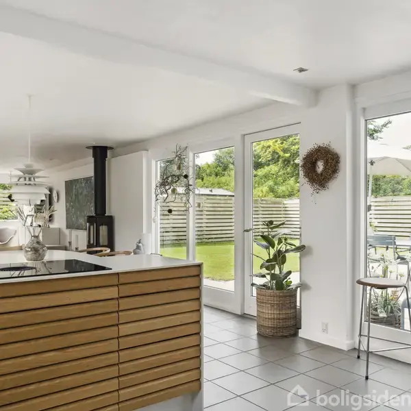 Køkkenøen står centralt, stilrent med trælameller, omgivet af en lys stue med vinduer og udsigt til en terrasse med borde og stole samt grønne planter udenfor.