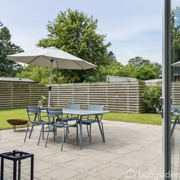 En terrasse med bord og fire stole under en parasol. Haven er omkranset af hegn med grøn beplantning og enkelte træer i baggrunden. Sollys skinner gennem åbne døre.