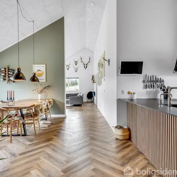 Spisebord med stole står i et åbent køkken-alrum. På væggen hænger dekorative hjortegevirer. Lyset strømmer ind fra et stort vindue, og køkkenet har moderne apparater.