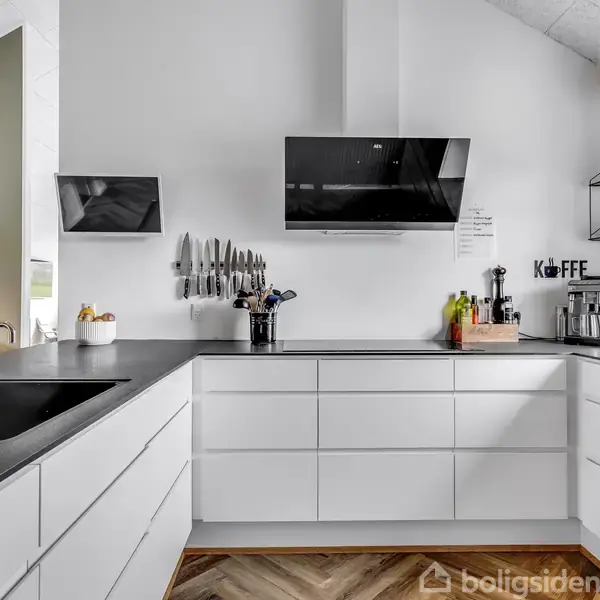 Moderne hvidt køkken med L-formet bordplade. Knive hænger på væggen, og en kaffemaskine står ved siden af teksten "Kaffe". Trægulv og minimalistisk udsmykning præger rummet.