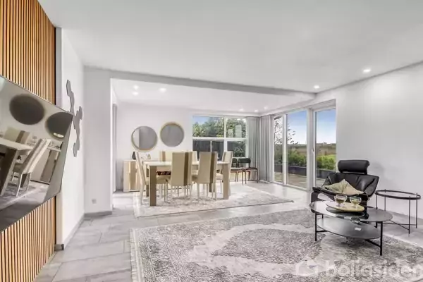 Stuen har moderne møbler med en lænestol og sofabord i forgrunden. Et spisebord med stole står nær vinduerne, der åbner mod en terrasse med udsigt.