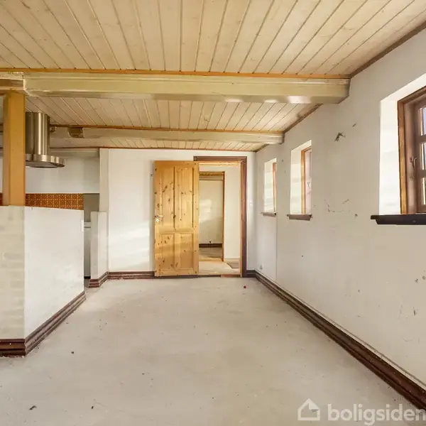 Et tomt rum med træloft har vinduer langs væggen og en åben dør mod et tilstødende område. Køkkenet ses til venstre med hvide vægge og gule søjler.