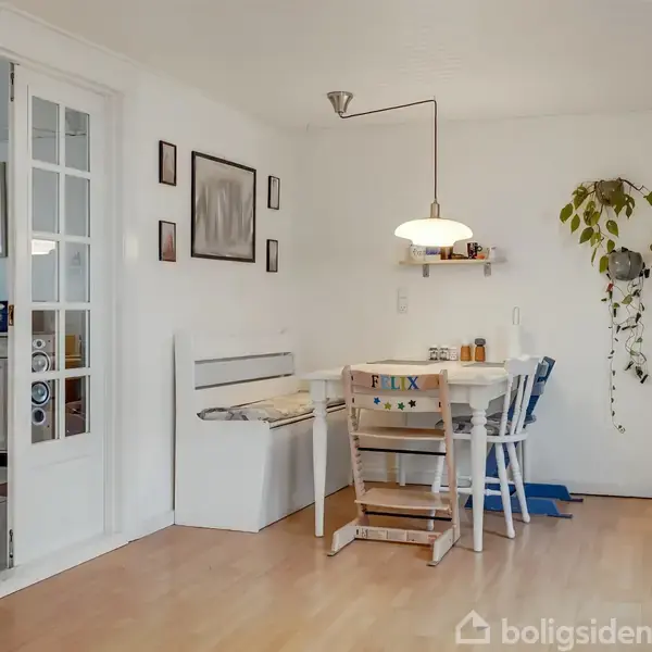 Spisebord med stole og en bænk står i et lyst rum med trægulve. En lampe hænger over bordet. En plante hænger fra væggen. Tekst på stolen: "FELIX".