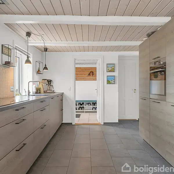 Køkken med lyse træskabe og en lang bordplade, hvor køkkenapparater er placeret. To pendellamper hænger over bordpladen. Lysindtag fra vinduer i et moderne miljø.