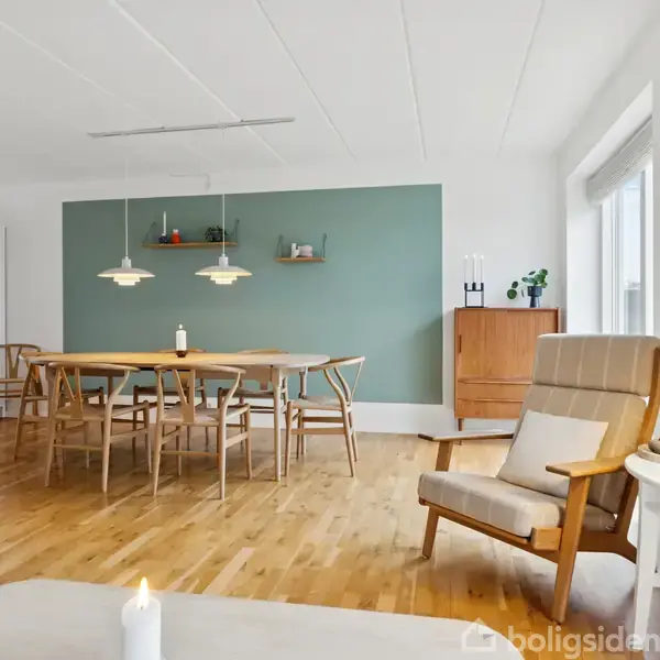 Stue med spisebord og stole, oplyst af hængelamper foran en grøn væg. Ved siden af står en lænestol og et lille bord med lampe. Store vinduer giver naturligt lys.