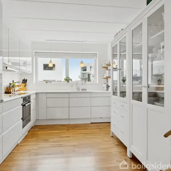 Et hvidt køkken med trægulv indeholder en indbygget ovn og over- og underskabe. Et vitrineskab står til højre. Fra vinduet ses hængende lamper og planter.