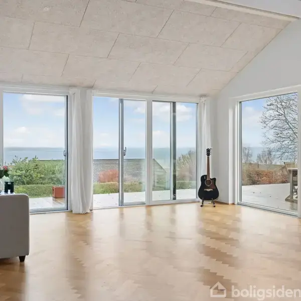 Lyst rum med parketgulv og store vinduespartier ud mod en terrasse. En sofa står til venstre, en akustisk guitar står ved vinduet, og udenfor ses havudsigt.