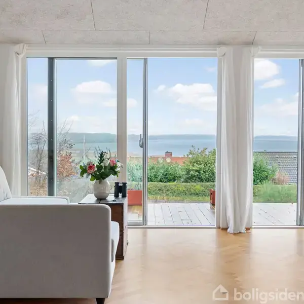 En hvid sofa står i en lys stue med store glasdøre. Dørene åbner ud til en terrasse med udsigt over havet og et grønt landskab.