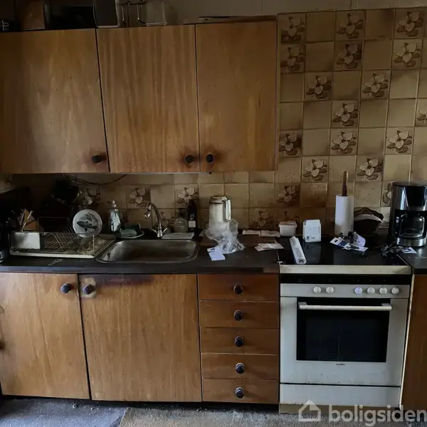 Køkken med træskabe, vask og komfur, omgivet af flisevægge med mønster. Køkkenredskaber står på bordet, og til højre er der en kaffemaskine. En stue ses i baggrunden.