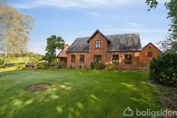 Et rødt murstenshus med skråt tag står i en grøn have med træer og buske omkring. Sollys skinner på græsplænen, der omkranser huset.