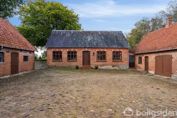 En gammel murstensbygning står stille på en brostensbelagt gårdsplads, omgivet af to mindre bygninger med røde tegltage, i en landlig og frodig træbevokset baggrund.