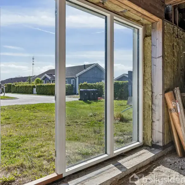 En stor skydedør står åben mod en græsplæne og en vej udenfor, med et hus i baggrunden. Indenfor ligger byggematerialer som en stige og et panel stablet op.