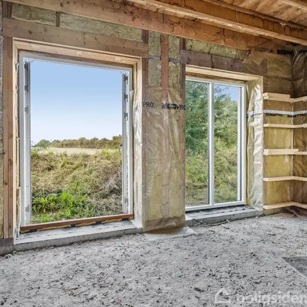 To store vinduer slipper lys ind i et ufærdigt rum med isolerede vægge, der åbner ud til en grøn have og en blå himmel. Golvet er råt beton.