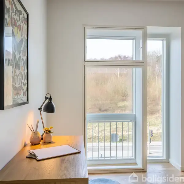 Et træskrivebord med en sort lampe og notesblok står ved et stort vindue. Udenfor ses et landskab med træer. På væggen hænger et billede.